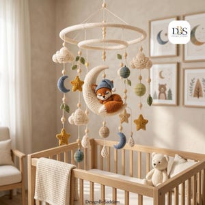 Op de afbeelding: Een gehaakte babymobiel met een centrale crèmekleurige halve maan met een slapende oranje vos met een blauwe muts. Hangende elementen zijn onder meer crèmekleurige wolken, gele sterren, blauwe en groene bollen en kleine blaadjes aan twee ringen.