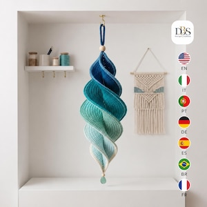 Puede incluir: Una decoración en espiral colgante hecha de hilo de ganchillo en un degradado ombré de azul, turquesa y verde claro. La pieza cuenta con una cuenta de madera en la parte superior e inferior, con un pequeño colgante de cristal verde en la base.