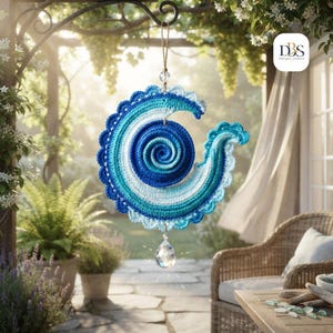May include: A circular crochet wall hanging featuring a spiral design in shades of dark blue, medium blue, light blue, and white. The piece has a scalloped edge and hangs from a string with a clear, faceted crystal drop suspended at the bottom.