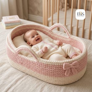 Könnte beinhalten: Ein Baby liegt in einem ovalen, handgehäkelten Stubenwagen mit abwechselnden horizontalen Streifen aus zartrosa und cremefarbenem Garn. Der Stubenwagen verfügt über zwei integrierte Griffe und eine dekorative Häkelschleife an der Vorderseite.