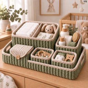 Può includere: Un set di cestini portaoggetti all'uncinetto color verde salvia e crema disposti su un vassoio abbinato. Il set include un grande cestino diviso contenente pannolini e un coniglietto di peluche, una scatola con coperchio, due piccoli contenitori quadrati con massaggiagengive a forma di orsetto in legno e un contenitore alto con articoli da toeletta.
