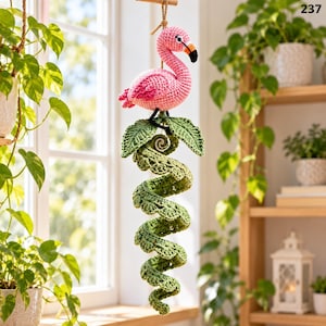May include: A hanging crochet decoration featuring a pink flamingo with a black and orange beak, perched atop a green spiraling vine with textured leaves. The entire piece is suspended by a thin brown cord, showcasing intricate yarn work.