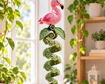 Puede incluir: Una decoración de ganchillo colgante que presenta un flamenco rosa con pico negro y naranja, posado sobre una enredadera verde en espiral con hojas texturizadas. La pieza cuelga de un cordel marrón fino, mostrando un intrincado trabajo de hilo.