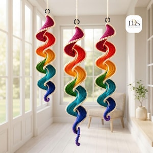May include: Three hanging spiral decorations featuring a vibrant rainbow gradient of magenta, red, orange, yellow, green, teal, blue, and purple yarn. Each spiral is edged with small white beads and suspended from a white cord with a black ring.