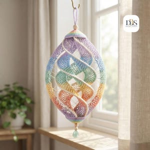 May include: A hanging spiral ornament crafted from pastel-colored crochet yarn in shades of lavender, blue, green, yellow, and peach. The structure features a central wooden bead at the top and bottom, with a small green teardrop charm at the base.
