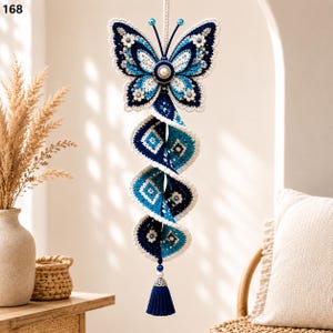 Op de afbeelding: Een hangende gehaakte decoratie met een vlinder aan de bovenkant met blauwe, witte en donkerblauwe vleugels, verbonden aan een spiraalvormige, meerlaagse gehaakte staart in bijpassende blauwtinten, afgewerkt met een donkerblauwe kwast onderaan.