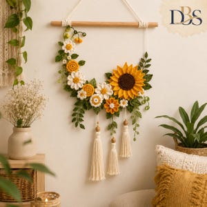 May include: A wall hanging featuring a crescent-shaped arrangement of crocheted sunflowers, daisies, and green leaves attached to a wooden dowel. Three cream-colored yarn tassels with wooden beads hang from the bottom of the floral display.