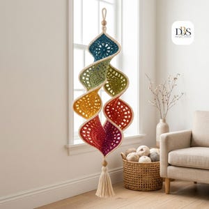 May include: A hanging spiral crochet decoration featuring a gradient of colors including teal, olive green, yellow, orange, red, and magenta. The piece is finished with cream-colored trim, a wooden bead at the top and bottom, and a cream-colored tassel.