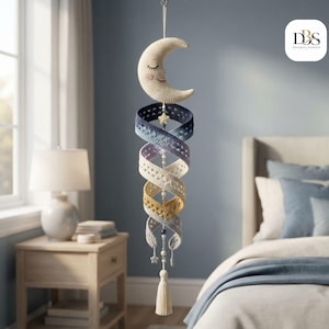 May include: A hanging crochet mobile featuring a cream-colored crescent moon with a stitched face at the top, followed by a spiraling, multi-colored ribbon in shades of dark blue, light blue, lavender, yellow, and grey, finished with a tassel.