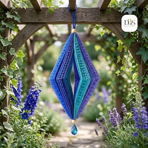 May include: A diamond-shaped hanging ornament crafted from crocheted fabric in shades of blue, teal, and purple. The structure features a central opening, a wooden bead at the top and bottom, and a blue teardrop-shaped crystal pendant at the base.