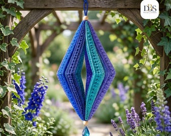 Puede incluir: Un adorno colgante en forma de diamante hecho de tela de ganchillo en tonos azul, turquesa y morado. La estructura presenta una abertura central, una cuenta de madera en la parte superior e inferior y un colgante de cristal azul en forma de gota.