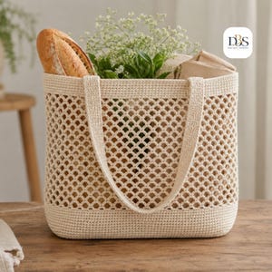 May include: A cream-colored crochet tote bag with a diamond-shaped openwork mesh pattern, a solid base, and two sturdy handles. The bag is filled with a crusty baguette, green foliage, and a folded brown paper item. The logo DBS is visible.