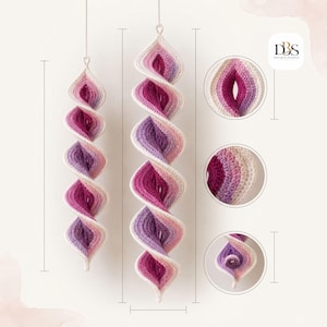 May include: Two hanging crochet spiral ornaments featuring a gradient color scheme of cream, light pink, magenta, and purple. The design includes a central twisted structure with circular detail insets showing the texture and the DBS logo.