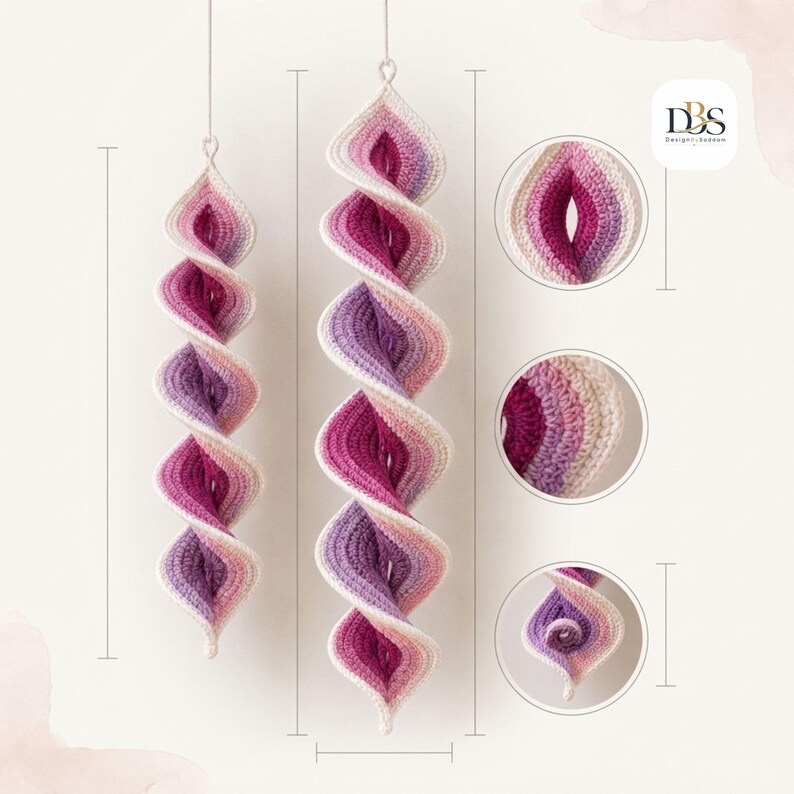 May include: Two hanging crochet spiral ornaments featuring a gradient color scheme of cream, light pink, magenta, and purple. The design includes a central twisted structure with circular detail insets showing the texture and the DBS logo.