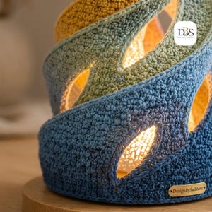 May include: A spiral-shaped crochet lamp shade featuring a gradient of blue, sage green, and yellow yarn. The textured fabric has open leaf-shaped cutouts that emit a warm glow. A small metal tag reads DesignBySaddam, and a logo reads DBS.