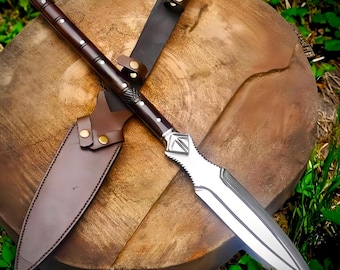 May include: A spear with a polished silver metal blade featuring a central ridge and a dark brown wooden shaft. The shaft is accented with small silver rivets and a textured metal collar. A brown leather sheath with brass snaps is included.