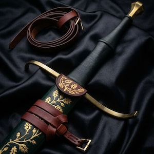 Peut inclure: Une épée avec une poignée gainée de cuir noir, un pommeau et une garde en laiton. Le fourreau vert foncé présente des motifs dorés de feuilles de chêne et de glands, fixés par des lanières en cuir marron avec une boucle en laiton.