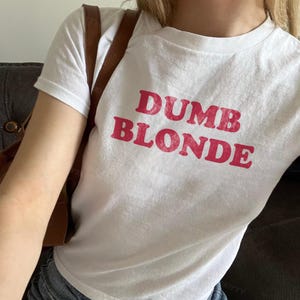 May include: A white short-sleeved t-shirt featuring the text DUMB BLONDE printed in a distressed red font across the chest. The shirt has a crew neckline and is worn with a brown shoulder bag strap visible over the shoulder.