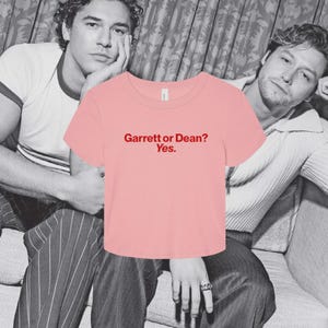 May include: A light pink short-sleeved crop top with a crew neckline. The center of the shirt features the text "Garrett or Dean? Yes." printed in a bold red font. The garment has a simple, clean design with a standard hemline and short sleeves.