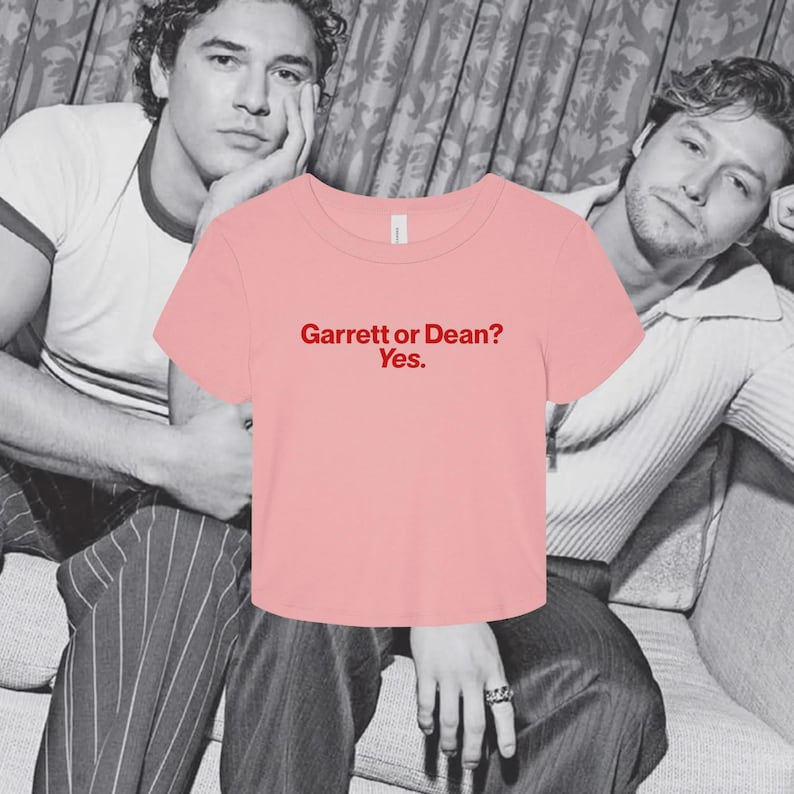 May include: A light pink short-sleeved crop top with a crew neckline. The center of the shirt features the text "Garrett or Dean? Yes." printed in a bold red font. The garment has a simple, clean design with a standard hemline and short sleeves.