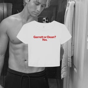 May include: A white short-sleeved crop top featuring the red text "Garrett or Dean? Yes." printed in a simple sans-serif font across the center of the chest. The garment has a round neckline and a straight hemline, constructed from a plain fabric.