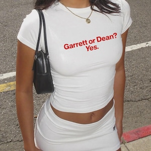 Puede incluir: Una camiseta blanca de manga corta y cuello redondo con el texto en rojo "Garrett or Dean? Yes." impreso en el pecho. La prenda se combina con una minifalda blanca a juego, un bolso de hombro negro y un collar de cadena dorada.
