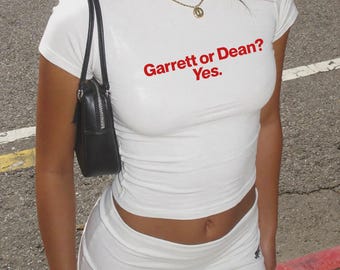 Pode incluir: Uma t-shirt branca de manga curta e gola redonda com o texto vermelho "Garrett or Dean? Yes." estampado no peito. A peça é combinada com uma minissaia branca a condizer, uma mala de ombro preta e um colar de corrente dourada.