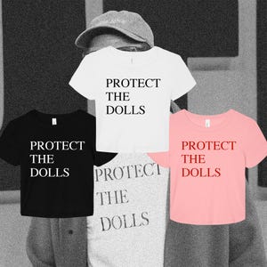 May include: A collection of four short-sleeved crop tops featuring the text PROTECT THE DOLLS. The shirts are available in white with black text, black with white text, light pink with red text, and a heather gray with dark gray text.