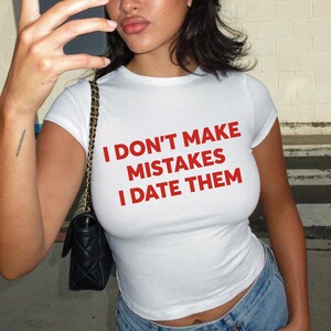 Puede incluir: Una camiseta blanca de manga corta con el texto en rojo I DON'T MAKE MISTAKES I DATE THEM impreso en el pecho. La camiseta es ajustada, tiene cuello redondo y se combina con un bolso de hombro negro con una cadena dorada.