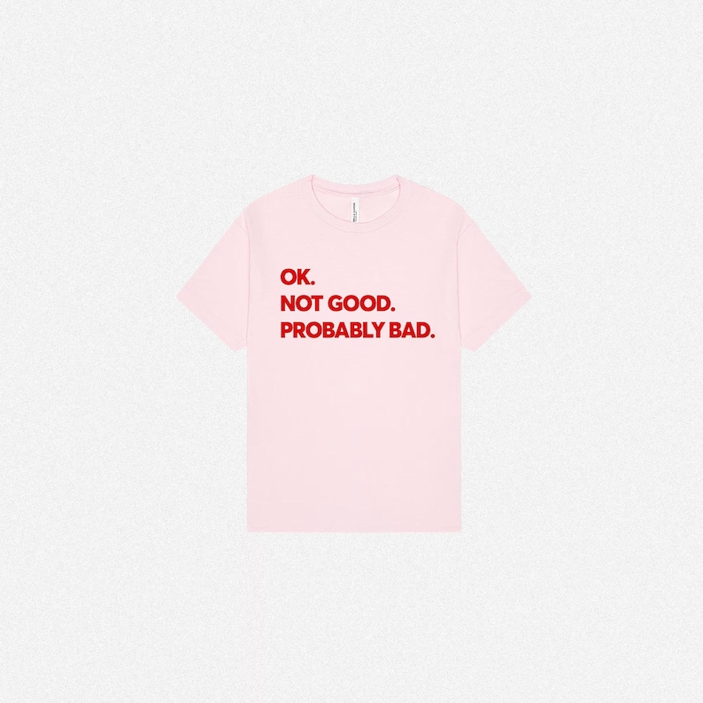 Pode incluir: Uma t-shirt de manga curta em rosa claro com o texto em letras vermelhas que diz: OK. NOT GOOD. PROBABLY BAD. A pe&ccedil;a tem um decote redondo e um corte padr&atilde;o, apresentando um design simples com tipografia em negrito centrada &agrave; frente.