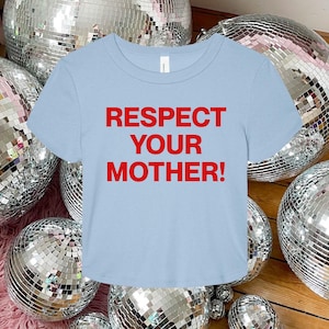 Op de afbeelding: Een lichtblauw t-shirt met korte mouwen en de tekst "RESPECT YOUR MOTHER!" in dikke, rode hoofdletters op het midden van de borst. Het kledingstuk heeft een ronde hals en een standaard pasvorm met een eenvoudig en strak ontwerp.