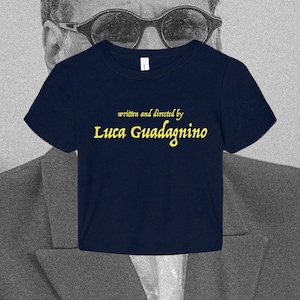 May include: A dark navy blue short-sleeved t-shirt featuring the yellow text "written and directed by Luca Guadagnino" printed across the center of the chest. The shirt has a crew neckline and a standard fit, showcasing clear, bold lettering.