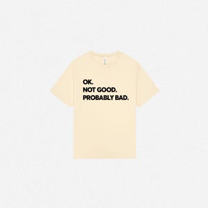 Pode incluir: Uma t-shirt de manga curta na cor creme com texto preto impresso no peito que diz: OK. NOT GOOD. PROBABLY BAD. A pe&ccedil;a tem um decote redondo padr&atilde;o e um corte relaxado, apresentada sobre um fundo neutro e liso.