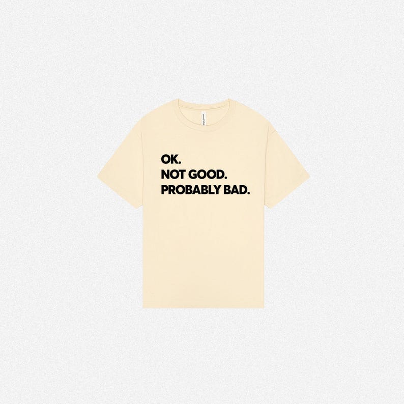 Pode incluir: Uma t-shirt de manga curta na cor creme com texto preto impresso no peito que diz: OK. NOT GOOD. PROBABLY BAD. A pe&ccedil;a tem um decote redondo padr&atilde;o e um corte relaxado, apresentada sobre um fundo neutro e liso.