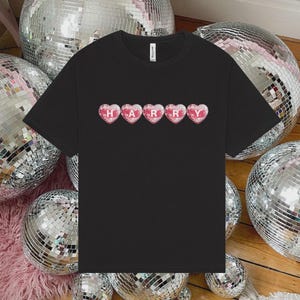 May include: A black short-sleeved t-shirt featuring a graphic design of five pink heart-shaped disco balls arranged in a horizontal row. Each heart contains a single white letter, spelling out the name HARRY across the center of the chest.