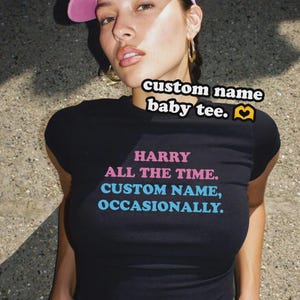 May include: A black short-sleeved baby tee featuring pink text that reads HARRY ALL THE TIME and blue text that reads CUSTOM NAME, OCCASIONALLY. The shirt has a crew neckline and a fitted silhouette, with additional text above reading custom name baby tee.