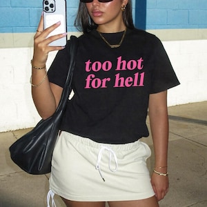 May include: A black short-sleeved t-shirt with the text "too hot for hell" printed in pink letters on the front. The shirt is paired with a cream-coloured mini skirt featuring an elastic waistband and white drawstring ties at the front.