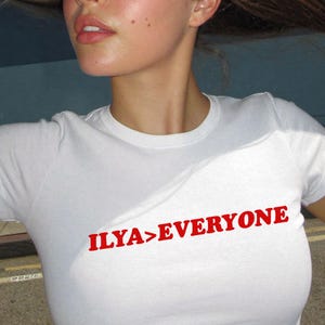 May include: A white short-sleeved t-shirt featuring the text ILYA>EVERYONE printed in a bold red font across the chest. The garment has a classic crew neckline and a fitted silhouette, providing a clean and simple look for casual daily wear.