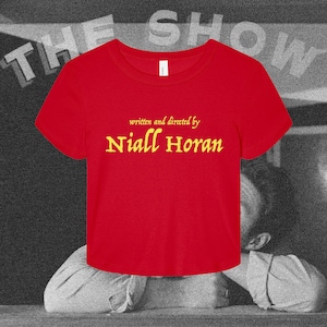 May include: A bright red short-sleeved t-shirt featuring the yellow text written and directed by Niall Horan printed on the front. The shirt has a round neckline and a cropped hemline, presenting a simple design with bold contrasting colors.