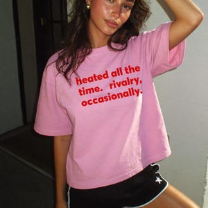 Pode incluir: Uma t-shirt de manga curta cor-de-rosa claro com o texto vermelho "heated all the time. rivalry, occasionally." estampado no peito. A camisola tem um decote redondo e um corte descontraído, combinada com calções desportivos pretos.