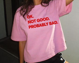 May include: A light pink short-sleeved t-shirt featuring the red text OK. NOT GOOD. PROBABLY BAD. printed on the front. The shirt has a crew neckline and a relaxed fit, paired with black athletic shorts that have a small white star logo on the leg.