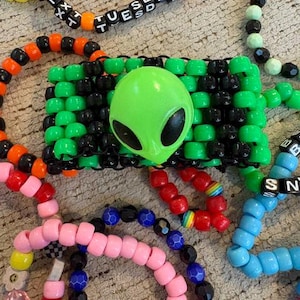 May include: A wide cuff bracelet made of neon green and black plastic pony beads featuring a large, central lime green alien head charm. The cuff is surrounded by several other beaded bracelets in various colors including pink, orange, yellow, and blue.