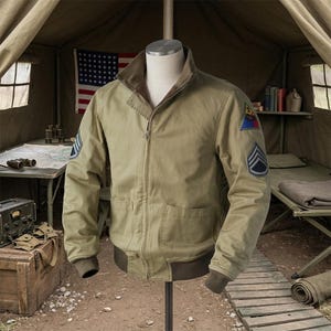 Könnte beinhalten: Eine beigefarbene Baumwolljacke mit durchgehendem Frontreißverschluss, Stehkragen und gerippten Bündchen. Die Jacke ist mit einem dreieckigen Aufnäher auf der linken Schulter und einem Chevron-Abzeichen an beiden Ärmeln versehen.