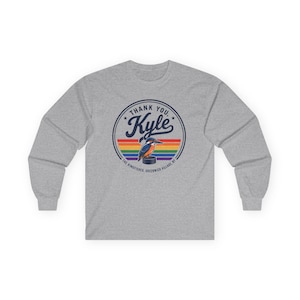 Pode incluir: Uma camisola de manga comprida cinzenta mesclada com um gráfico circular com o texto "THANK YOU, Kyle" acima de um guarda-rios pousado num disco de hóquei, sobre um fundo de riscas horizontais arco-íris e o texto "THE KINGFISHER, GREENWICH VILLAGE, NY".