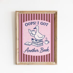Puede incluir: Una lámina enmarcada que muestra un pato blanco sentado sobre una pila de libros leyendo, con una taza de café al lado. El texto dice OOPS! I GOT Another Book. El fondo tiene rayas verticales marrones y rosas con un centro festoneado.