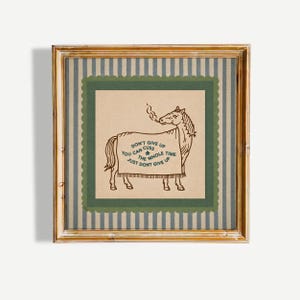 May include: A square framed wall art piece featuring a tan background with a brown line drawing of a horse wearing a blanket. The blanket text reads: DON'T GIVE UP YOU CAN CUSS THE WHOLE TIME JUST DONT GIVE UP. The frame has a grey and white striped border.