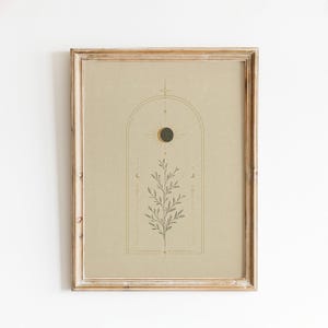 May include: A framed wall art print featuring a tan background with a centered dark green botanical illustration of a leafy branch enclosed within a thin arched line drawing. Above the branch is a dark circle representing a moon phase.