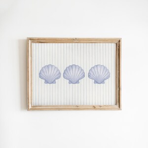 May include: A rectangular wooden frame containing a print of three light purple scallop shells arranged horizontally. The background of the print features a pattern of thin, vertical light gray stripes on a white surface.