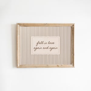 Peut inclure: Un cadre en bois rectangulaire contenant une impression avec un fond à rayures verticales beige et marron. Au centre, une étiquette rectangulaire à bords festonnés porte le texte cursif marron foncé : fall in love again and again.