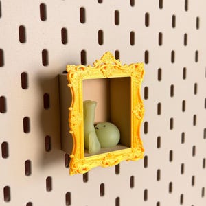 May include: A bright yellow, ornate, rectangular picture frame shelf mounted on a pegboard. Inside the frame sits a small, smooth, pale green, rounded figurine with a simple facial expression, positioned as if resting or reclining.